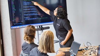 Eine junge Frau erklärt auf einem großen Screen anderen Frauen einen Computer Code