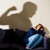 Eine Frau hält sich auf dem Sofa schützend die Hände über den Kopf. Als Schatten sieht man einen Mann, der die Faust hochhebt.