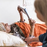 Eine Frau sitzt am Bett einer pflegebedürftigen Frau, die ein Buch liest (Symbolbild)