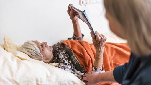 Eine Frau sitzt am Bett einer pflegebedürftigen Frau, die ein Buch liest (Symbolbild)