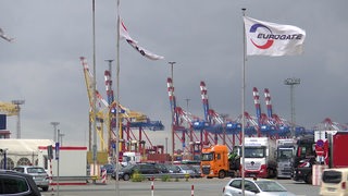 Im Hintergrund sind Containerbrücken von einem Hafen sichtbar. Davor sind Autos und Lkws zu sehen und Fahnen mit dem Schriftzug "Eurogate".