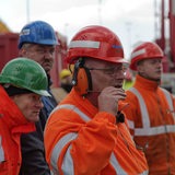 Hafenarbeiter und Lademeister mit Helmen stehen im Hafen, im Hintergrund Container und ein Ladekran.