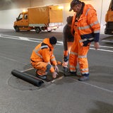 Bauarbeiter stehen vor Baufarzeugen in einem leeren Tunnel und arbeiten am Boden.