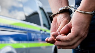 Einen Person steht in Handschellen gefesselt neben einem Polizeifahrzeug. 