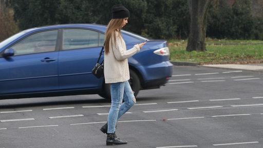 Junge Frau läuft unachtsam und ohne zu gucken mit Handy über die Straße. (Symbolbild)