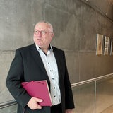Ein älterer Mann mit grauen Haaren und roter Brille steht vor einer grauen Wand. 