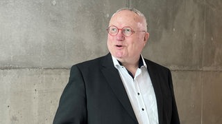 Ein älterer Mann mit grauen Haaren und roter Brille steht vor einer grauen Wand. 