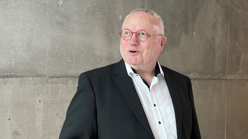 Ein älterer Mann mit grauen Haaren und roter Brille steht vor einer grauen Wand. 