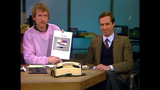 Moderator Michael Geyer hält im Gespräch mit Fernsehdirektor Hans-Werner Conrad ein Bild mit einem Testbild hoch.