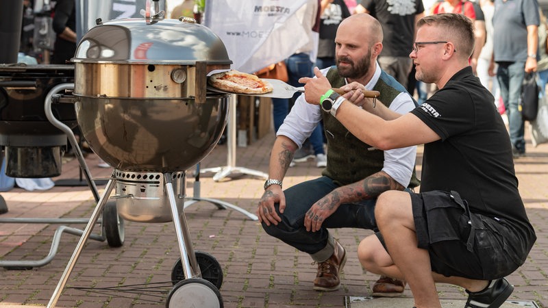 Zwei Männer knien vor einem Pizzaoffen, einer hat einen Pizzaschieber in der Hand.