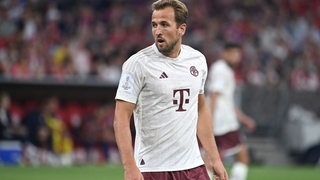 Fußballspieler Harry Kane steht in einem Trikot von Bayern München auf einem Fußballplatz.