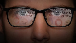 Nahaufnahme der Augenpartie eines Mannes, in dessen Brillengläsern sich Hasskommentare spiegeln.