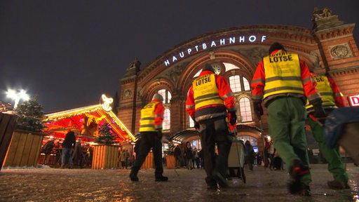 Vier Männer mit gelben Warnwesten vor dem erleuchteten Bremer Hauptbahnhof.