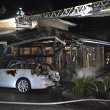 Ein Feuerwehrmann blickt auf ein abgebranntes Haus, vor dem ein stark beschädigtes Auto steht.