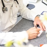 Eine Person mit Arztkittel und Stetoskop sitzt an einem Schreibtisch und füllt ein Rezept aus.