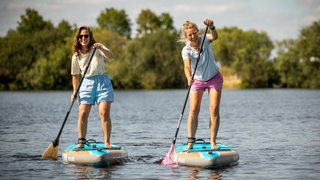 Zwei Frauen stehen lachend aud Paddelbrettern mit Paddeln in der Hand und fahren über einen See in der Sonne.