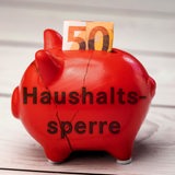 Ein rotes Sparschwein mit Rissen und der Aufschrift "Haushaltssperre" steht auf einem Tisch