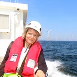 Eine Frau mit Schwimmweste und Helm steht auf einem Schiff vor einem Windpark.