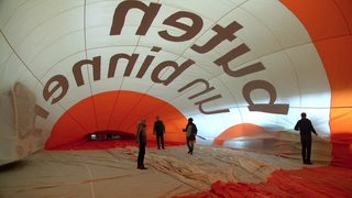 Personen stehen in einem zur Inspektion am Boden aufgeblasenden Heißluftballon. Der Ballon ist orange und weiß und trägt den Schriftzug "buten un binnen".