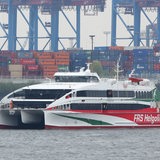 Ein Katamaran fährt in einem Hafen. Im Hintergrund sind Container zu sehen. 