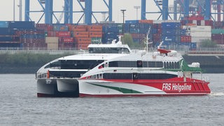 Ein Katamaran fährt in einem Hafen. Im Hintergrund sind Container zu sehen. 
