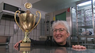 Die Europameisterin im Plattenlegen, Henrike Seliger, kniet neben ihrem Pokal in ihrer Cateringküche.