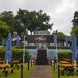 Bänke, Tische und Regenschirme stehen vor einem Restaurant mit dem Namen Ahoi auf grünem Rasen.