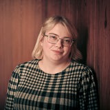 Eine junge Frau mit blonden Haaren lehnt an einer holzvertäfelten Wand und schaut in die Kamera.