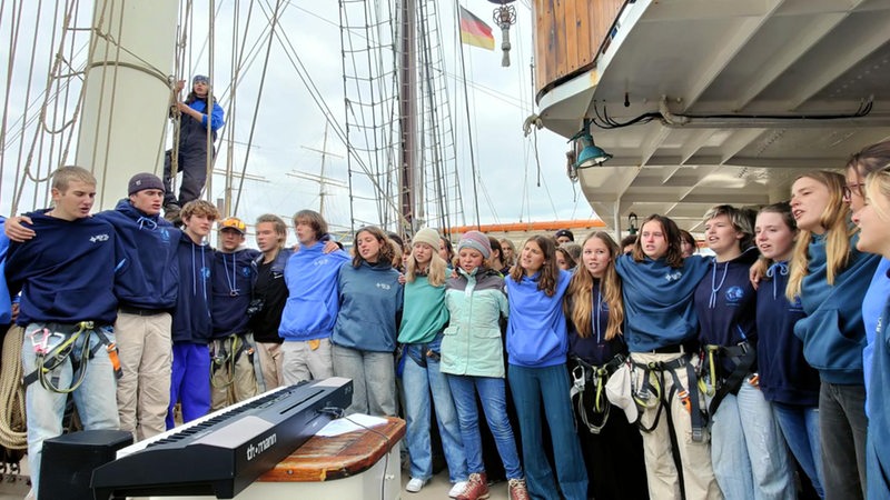 Mehrere junge Leute stehen Arm in Arm auf einem Segelschiff.