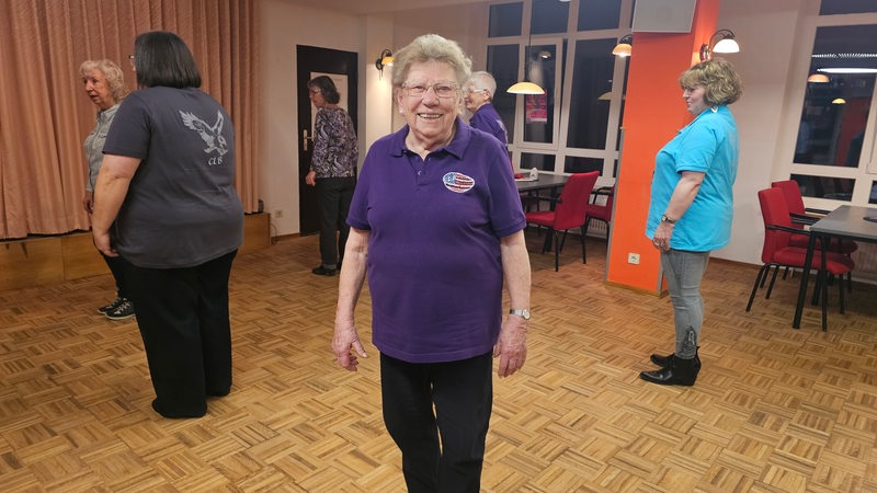 Hilde Tscharntke steht mit einem lilafarbenen T-Shirt auf einer Tanzfläche und lächelt 