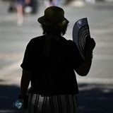 Eine ältere Frau mit Sonnenhut fächert sich im Schatten eines Baumes in der Stadt Luft zu.