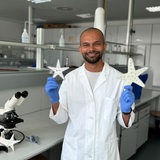 Doktorand Raman lächelt und hält zwei seiner Prototypen in den Händen, die beide die Form eines Seesterns haben. 