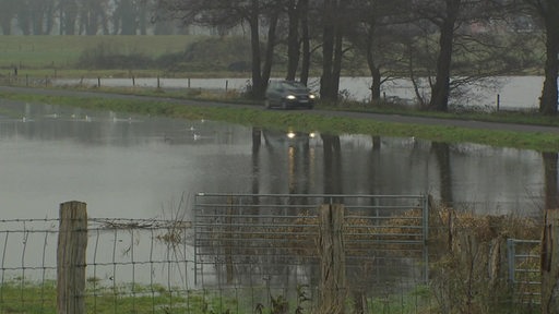 Es ist eine überschwemmte Landschaft zu sehen. In der Mitte fährt ein Auto über einen Damm.