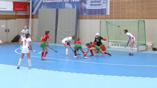 Zwei Damenmannschaften spielen in einer Turnhalle gegeneinander Hockey.