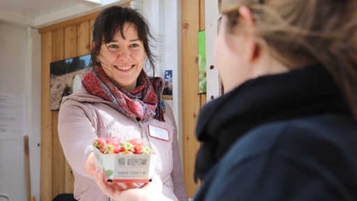 Eine Frau in einem Verkaufsstand überreicht einer Kundin eine Schale Erdbeeren.