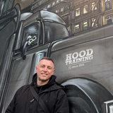 Ein junger Mann in einer schwarzen Jacke steht lächelnd vor einem Graffiti, das einen großen Truck zeigt, aus dessen Beifahrerfenster ein Waschbär schaut.