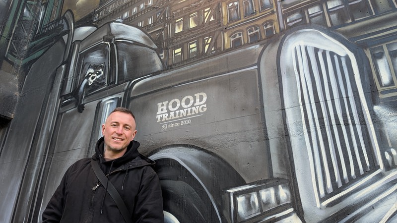 Ein junger Mann in einer schwarzen Jacke steht lächelnd vor einem Graffiti, das einen großen Truck zeigt, aus dessen Beifahrerfenster ein Waschbär schaut.