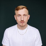 Hubertus Koch steht im weißen T-Shirt vor einer dunklen Wand und schaut in die Kamera