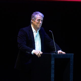 Starschauspieler Hugh Grant steht vor einem Pult und spricht in ein Mikrofon