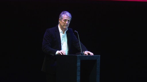 Der Schauspieler Hugh Grant steht vor schwarzem Hintergrund am Pult auf der Bühne des Goethe Theaters in Bremen