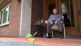 Eine ältere Dame sitzt auf einem Gartenstuhl vor ihrem Haus.