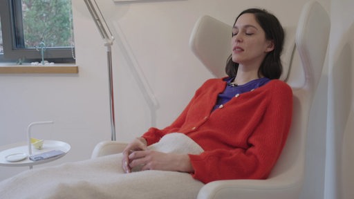 Die Reporterin Sophie Labitzke liegt während einer Hypnose in einem weißen Stuhl, sie trägt einen roten Cardigan und hat die Augen geschlossen