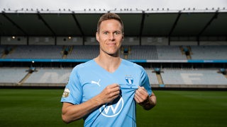Niklas Moisander strahlt bei seiner Vorstellung in Malmö im hellblauen Trikot in die Kamera und deutet auf das Wappen über der Brust.