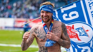 Rostocks Jan Löhmannsröben feiert nach dem Aufstieg oberkörperfrei mit einem Vereinsschal um die Stirn gebunden und einer Hansa-Rostock-Flagge.
