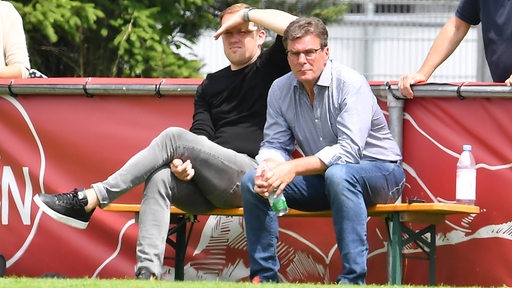 Olaf Rebbe und Dieter Hecking sitzen beim Training auf einer Bierbank und schauen gespannt zu. 