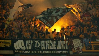 Fans von Dynamo Dresden zünden Pyrotechnik im Berliner Olympiastadion. Vor dem Block hängt ein Banner mit der Aufschrift "Ostdeutsche Jungs - reiselustig & erlebnisorientiert". 