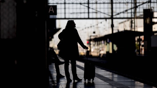 Eine Frau mit einem Rollkoffer wartet an einem Bahnsteig auf einen Zug.