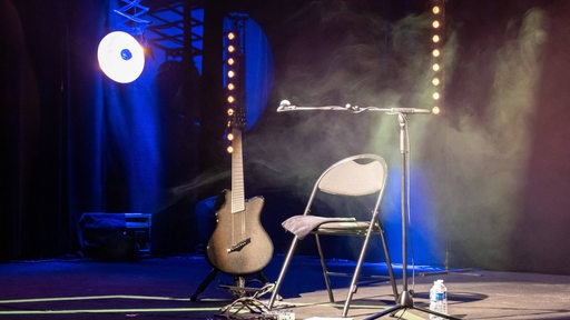 Ein leerer Stuhl auf Bühne im Scheinwerferlicht, ein Bass und Mikrofon stehen daneben