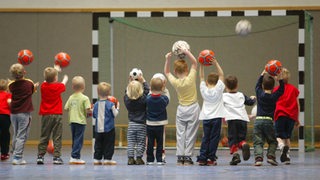 Kinder werfen in einer Sporthalle mit Bällen auf ein Handball-Tor.