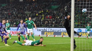 Werder-Spieler Milos Veljkovic liegt nach seinem Hackenschuss auf dem Boden und schaut zu, wie sich der Aue-Torwart vergeblich nach dem Ball streckt.